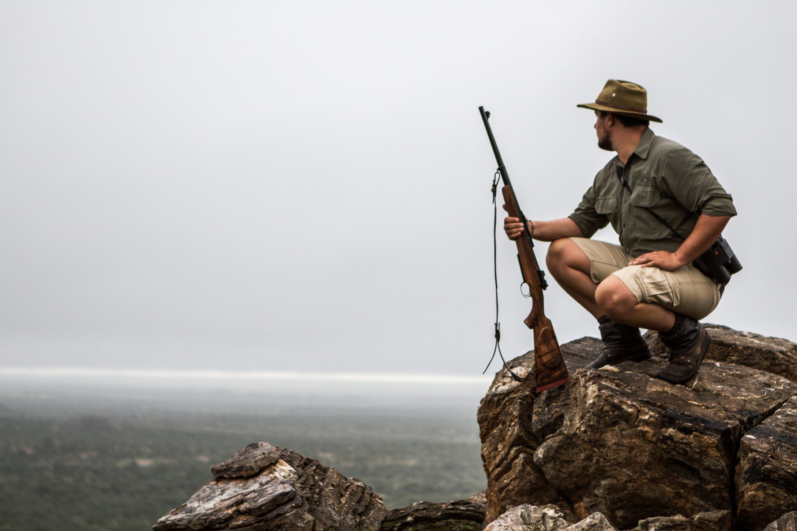 Man crouched on boulder with a rifle whilst looking over landscape