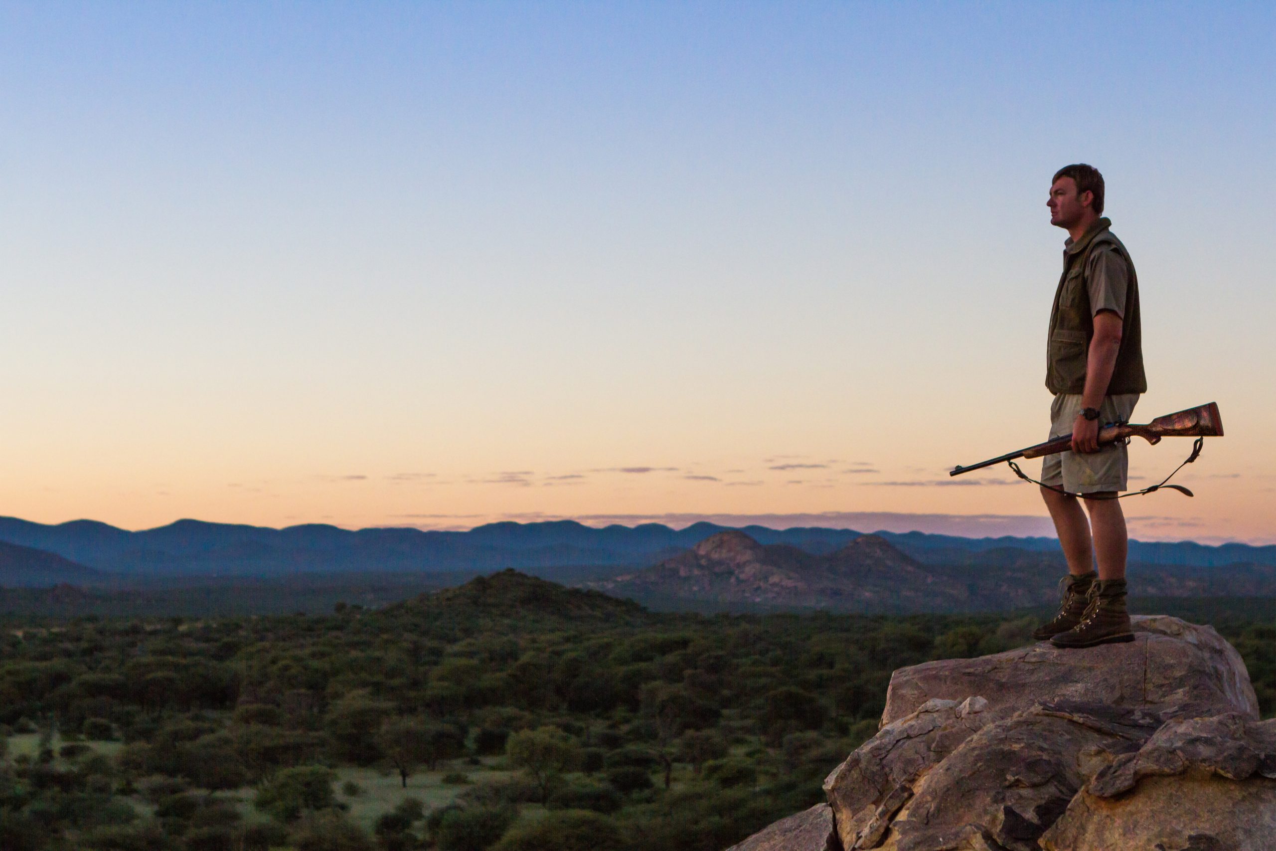 Man standing on a boulder looking out over the horizon with a rifle in his hand